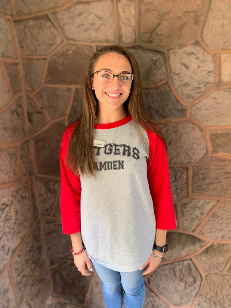 Katherine Conway, with a slight tan, medium brown hair, and black framed glasses, stands in front of the brick of the Admissions Building in a Rutgers—Camden baseball tee.